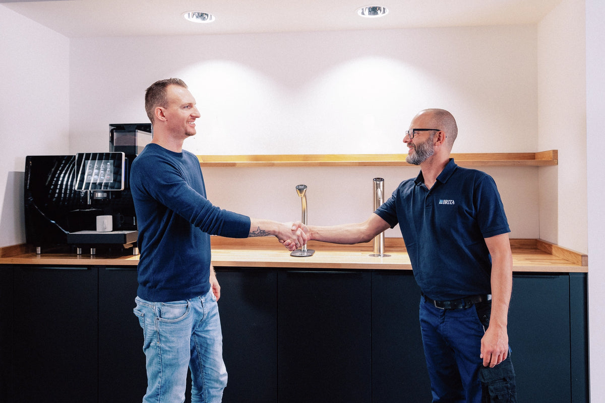 Two men shaking hands in a modern kitchen setting with water dispensers in the back.