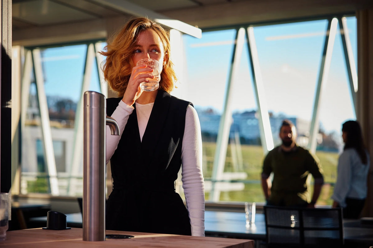A woman standing in front of an BRITA I-Tap Dispenser, drinking water from a glass in an office setting. 