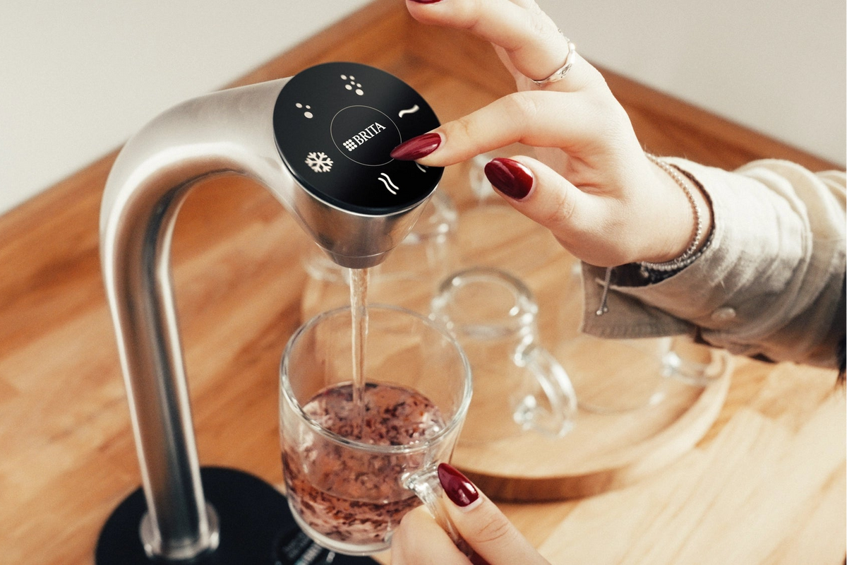 Woman using a BRITA Extra C-Tap to pour hot water into a cup on a wooden surface.